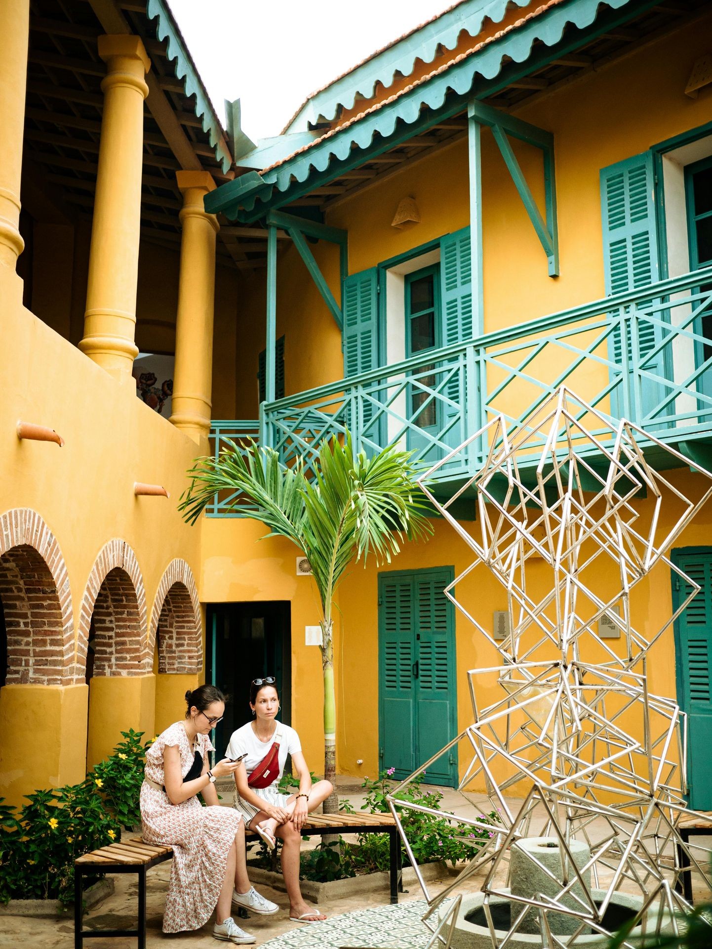 Two people seated in a courtyard with a yellow building, green shutters, and metal sculpture.